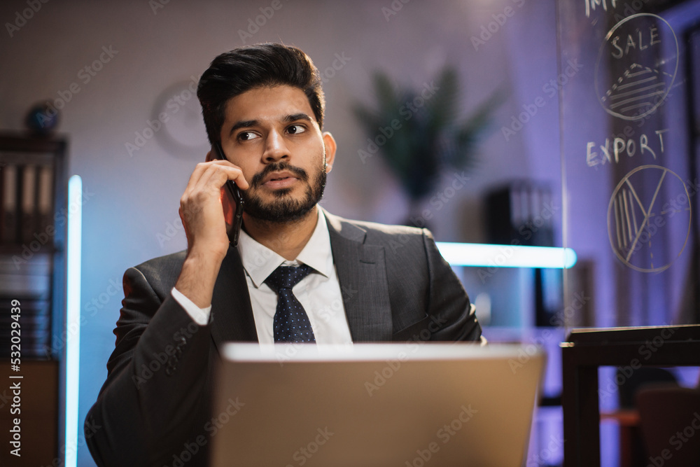 Close up portrait of handsome concentrated hardworking bearded indian businessman or manager in suit having business conversation with smart phone and laptop computer in evening office.