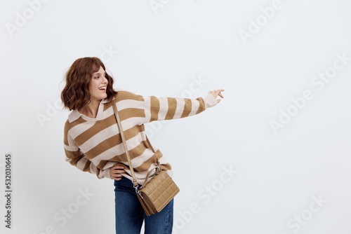 Fototapeta Naklejka Na Ścianę i Meble -  a pretty sweet woman stands on a light background in a striped sweater and a bag on her shoulder, leaned forward, pushing her hand away from her, pressing the other to her stomach