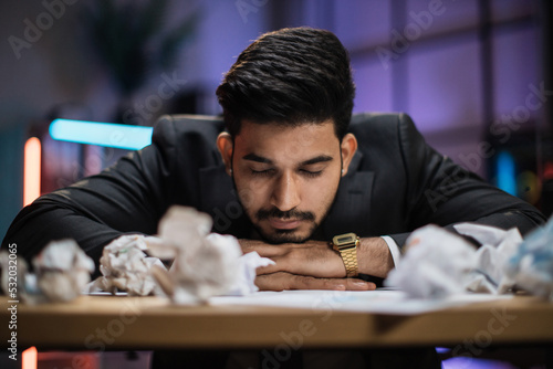 Portrait of tired overworked bearded arab businessman in formal suit, sleeping on desk with sheets of crumpled papers in eveninng office. photo