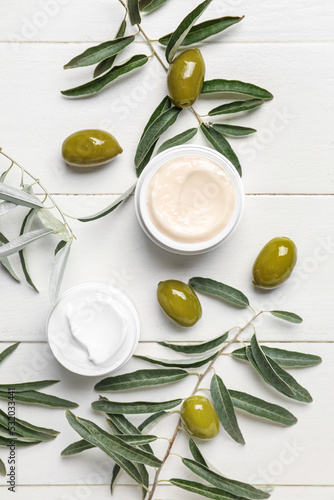 Jars of cream with green olives and plant branches on white wooden background