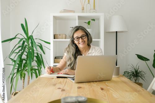 Portrait of adult woman working from home and looking at the camera, she is sitting at her desk with laptop open next to her and smiling to camera