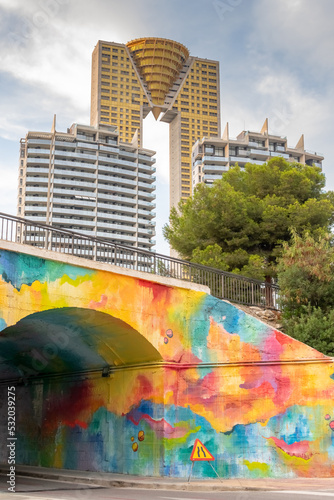 Intempo skyscraper and towers of Coblanca Murtal, trees and a cloudy sky in the background shot from C. Murtal under the colorful bridge of Villa Joiosa Avenue. photo