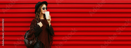 Portrait of beautiful young woman with cup of coffee wearing brown knitted poncho, black round hat on red background © rohappy