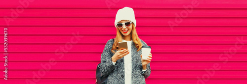 Portrait of stylish smiling woman with smartphone wearing white knitted hat, gray coat jacket on pink background © rohappy