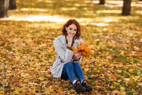 Smiling caucasian millennial red-haired woman in raincoat with yellow leaves in hands, sits on ground in park