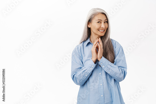 Smiling old asian lady, senior woman scheming, has plan, thinking of something with cunning, coy smile, steeple fingers, standing over white background