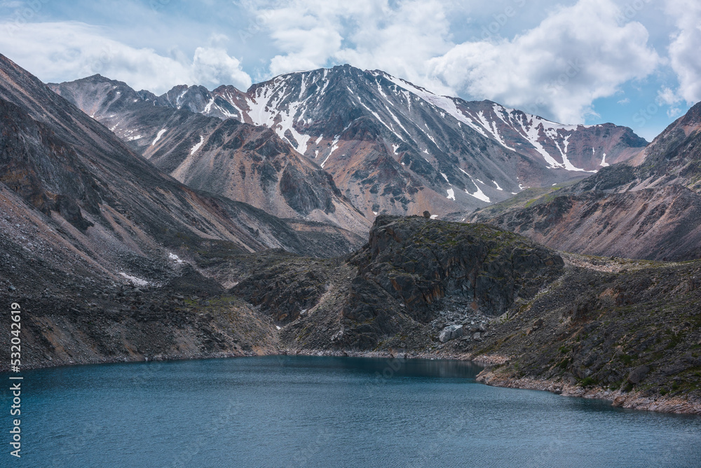 Obraz premium Deep mountain lake of phantom blue color among sharp rocks and high mountains in changeable weather. Wonderful dramatic view to deep blue mountain lake and large sunlit rocky cliff under cloudy sky.