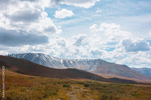 Motley autumn landscape with hills on high plateau and sunlit snowy mountain range under dramatic cloudy sky. Vivid autumn colors in mountains. Sunlight and shadows of clouds in changeable weather.