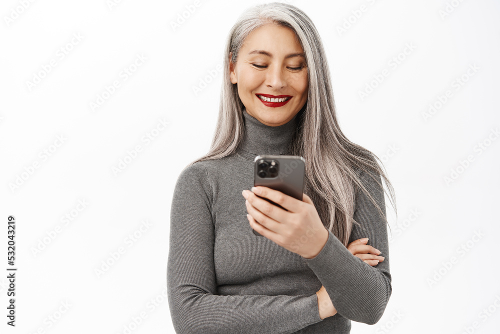 Portrait of beautiful middle aged asian woman holding smartphone and smiling at camera, using mobile app, standing over white background Portrait of beautiful middle aged asian woman holding smartphone and smiling at camera, using mobile app, standing over white background