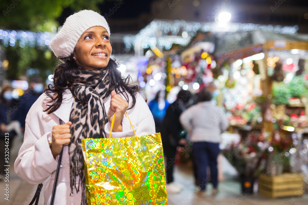 Fototapeta premium Smiling Latin American woman holding a gift package in her hands came to an open-air Christmas fair