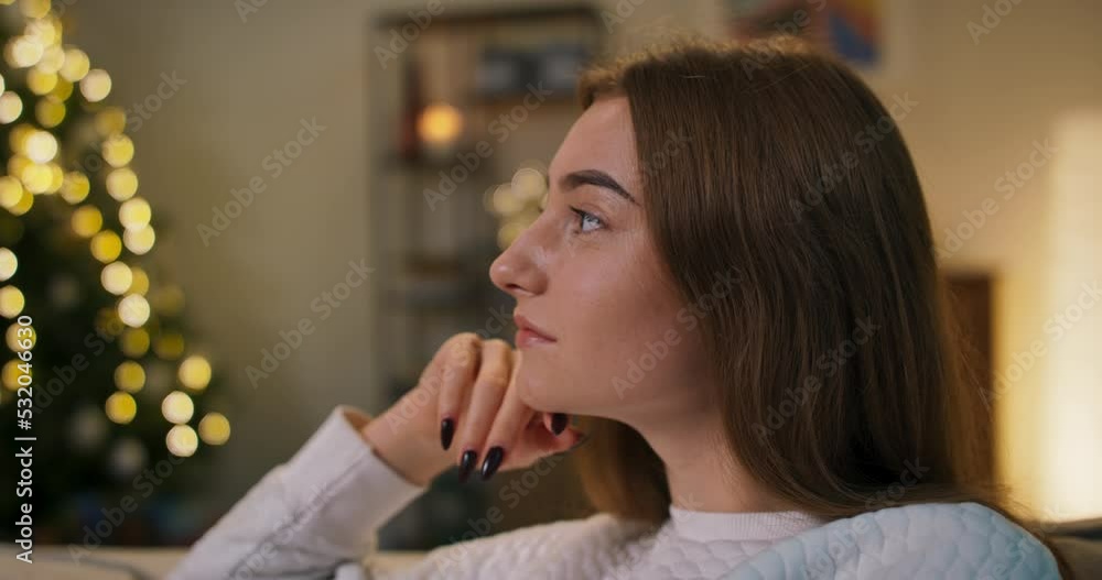 Close-up of girl with long hair. The girl rested head on hand and looked into the distance. She has black nails and white sweater. The girl dreams of new journey. Christmas tree flickers in the room. Close-up of girl with long hair. The girl rested head on hand and looked into the distance. She has black nails and white sweater. The girl dreams of new journey. Christmas tree flickers in the room.