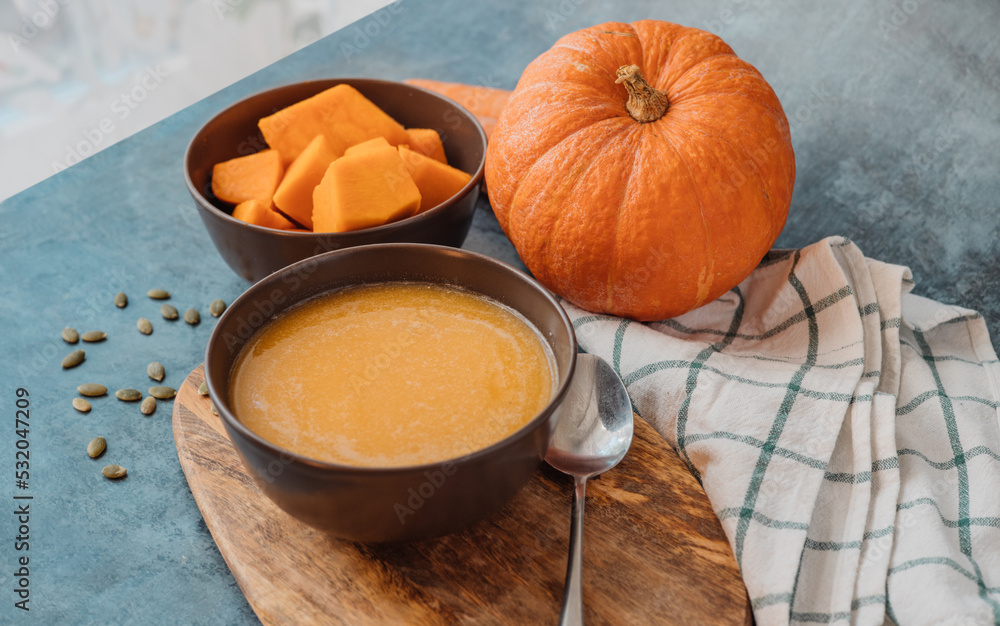 Pumpkin cream soup in a bowl with a spoon on wooden board on the table next to pumpkin whole and cut into pieces. Healthy, vegetarian food. Diet. Home cooking. Thanksgiving day. Halloween. Pumpkin cream soup in a bowl with a spoon on wooden board on the table next to pumpkin whole and cut into pieces. Healthy, vegetarian food. Diet. Home cooking. Thanksgiving day. Halloween.