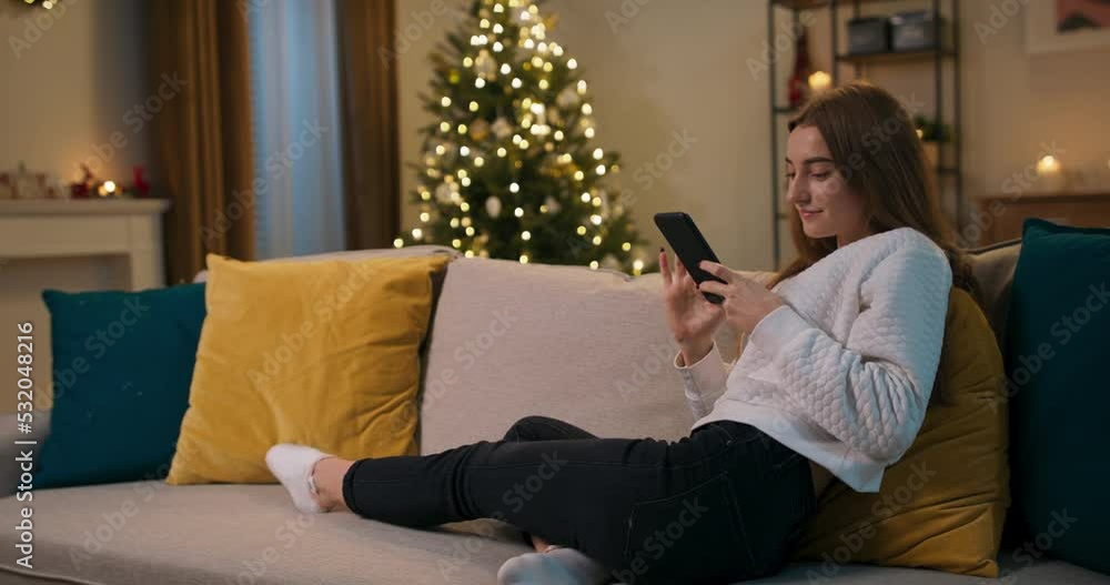 Winter evening. After work, the girl sits on couch in the living room. The beauty holds phone in hands, looks at funny photos and smiles. The room is cozy, shimmering Christmas tree. Winter evening. After work, the girl sits on couch in the living room. The beauty holds phone in hands, looks at funny photos and smiles. The room is cozy, shimmering Christmas tree.