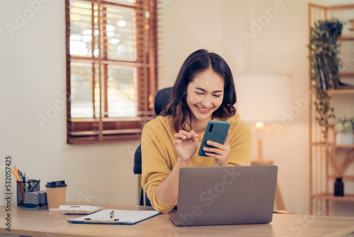 Attractive brunette Asian woman using mobile phone, smile and happy while sitting at working desk in modern office.