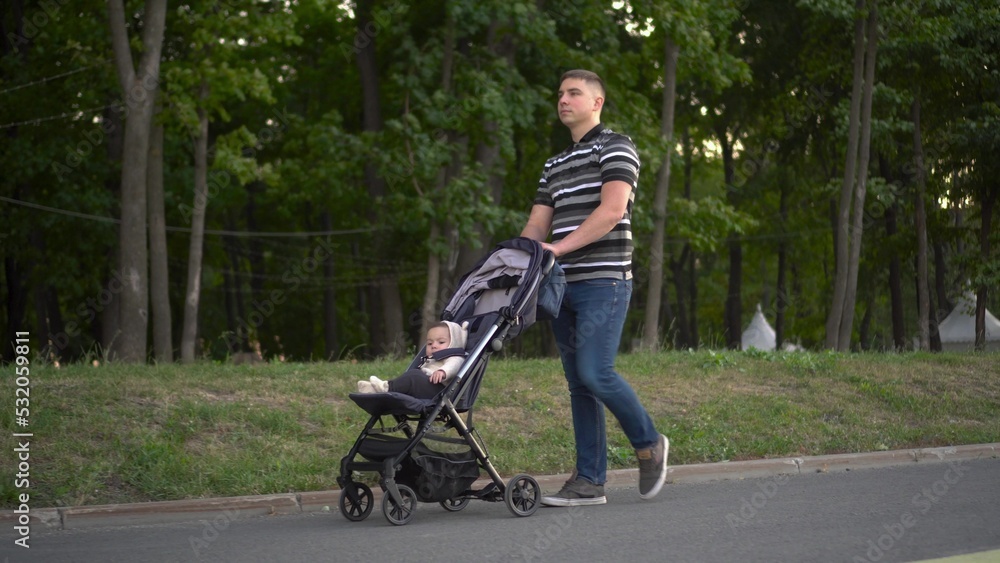 A young man is carrying a baby in a stroller through the park. Father and son are walking in nature.