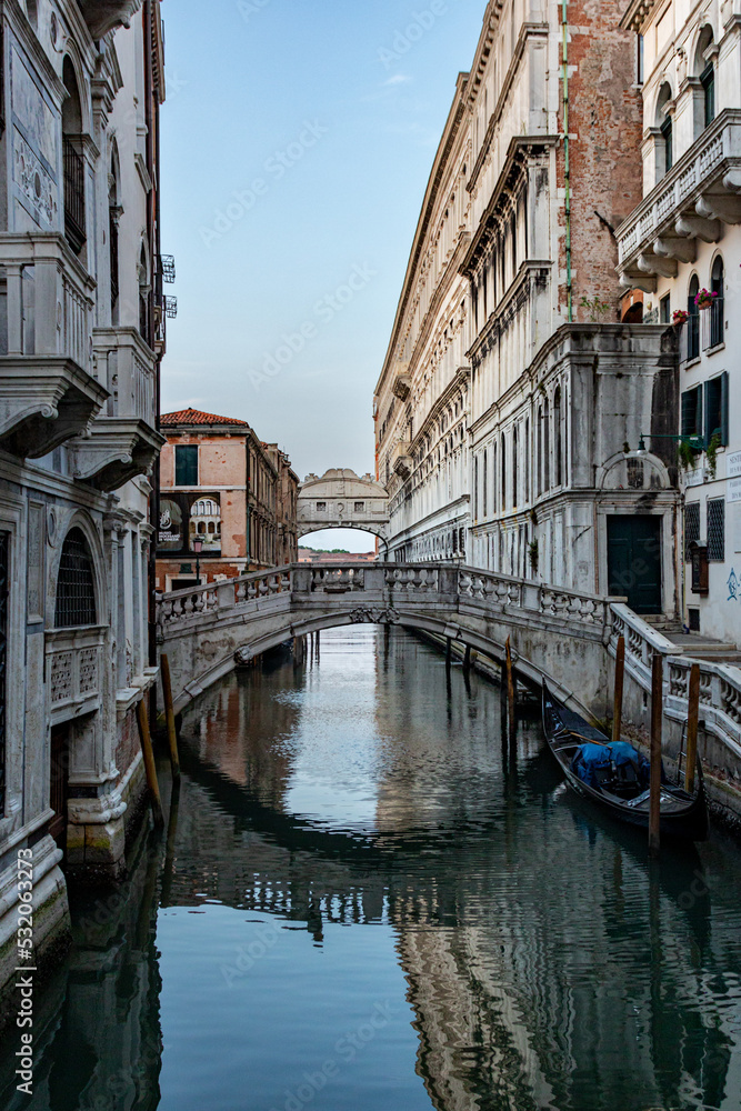 Narrow canal with medieval buildings, and Bridge of Sighs reflected in calm water  in Venice, Italy on sunny day.