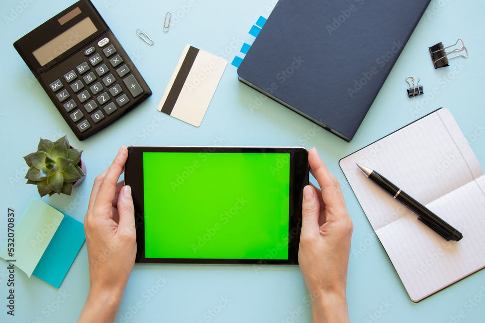 Tablet computer with green screen and female hands on blue background. Top view of desktop with various office supplies. Flat lay, mock up. Tablet computer with green screen and female hands on blue background. Top view of desktop with various office supplies. Flat lay, mock up.