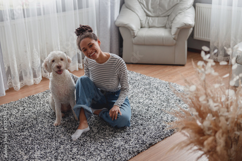 Mixed-race beautiful cheerful woman smiling in good mood sitting on carpet floor with dog