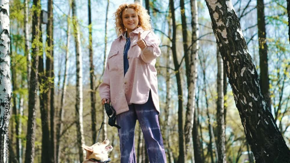 Young Caucasian Girl Running With Her Corgi Dog In The Park Show Thumbs Up In The Camera. - low angle Young Caucasian Girl Running With Her Corgi Dog In The Park Show Thumbs Up In The Camera. - low angle