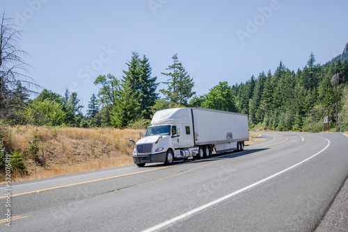 Clean white big rig semi truck with dry van semi trailer carry cargo running on the winding highway road in gorgeous Columbia Gorge National Reserve area © vit