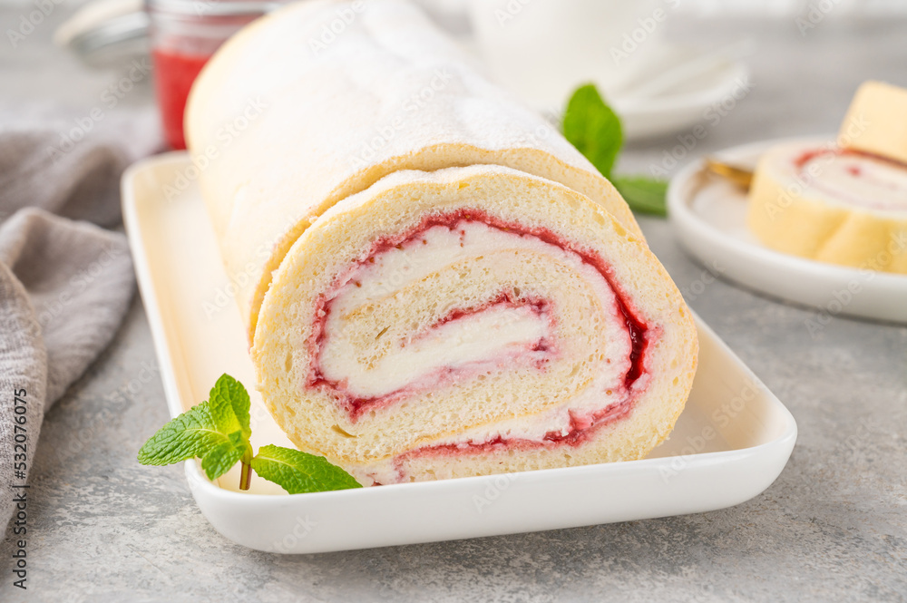 Sponge cake roll with strawberry jam and cream on a white plate on a gray background. Copy space. Sponge cake roll with strawberry jam and cream on a white plate on a gray background. Copy space.