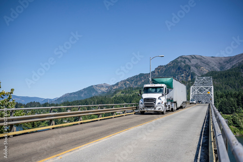 Local day cab big rig semi truck transporting cargo in bulk semi trailer driving on the truss arched bridge across the Columbia River In Columbia Gorge area