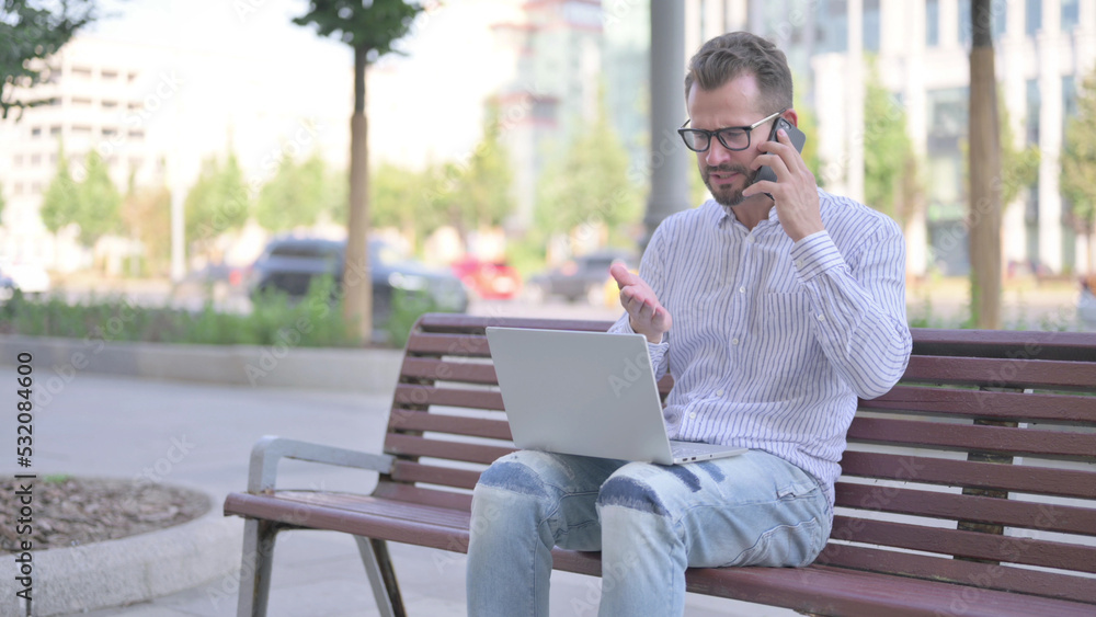 Fototapeta premium Angry Young Adult Man Talking on Phone and using Laptop while Sitting Outdoor on Bench