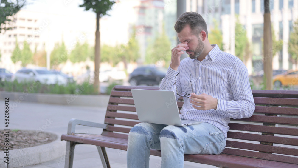 Young Adult Man with Headache Using Laptop while Sitting Outdoor on Bench Young Adult Man with Headache Using Laptop while Sitting Outdoor on Bench
