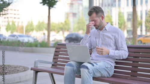 Young Adult Man with Headache Using Laptop while Sitting Outdoor on Bench © stockbakers