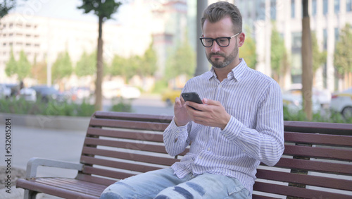 Young Adult Man Using Smartphone while Sitting Outdoor on Bench