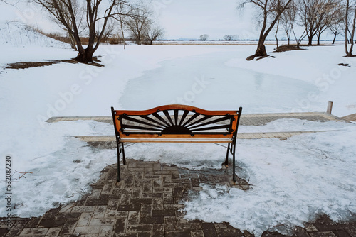 Cute bench near snow-covered river. Cozy winter walk in park.
