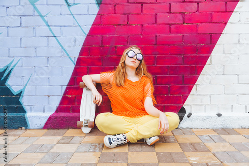 Valokuva An image of a beautiful 18-year-old girl laughing while sitting on a skateboard against a bright brick wall