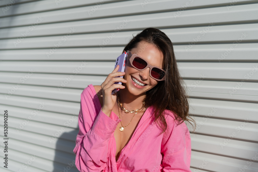 Young beautiful brunette woman in a pink shirt, neck jewelry, necklace, trendy sunglasses on the background of a light garage fence, calls the phone, laughs, smiles Young beautiful brunette woman in a pink shirt, neck jewelry, necklace, trendy sunglasses on the background of a light garage fence, calls the phone, laughs, smiles