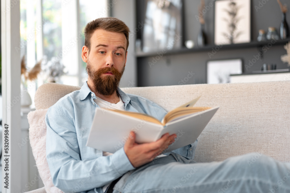 Young bearded handsome man reading book on couch at home