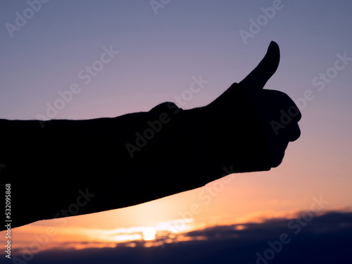 Silhouette of thumbs up gesture against warm and cool sunset sky. Expression of positive emotion through body language. Ok sign.