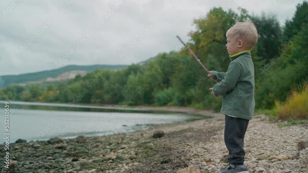 A three-year-old kid stands with a stick in his hands on the shore of a mountain lake. He swings his stick and wants to throw it into the water, but at the last moment he changed his mind A three-year-old kid stands with a stick in his hands on the shore of a mountain lake. He swings his stick and wants to throw it into the water, but at the last moment he changed his mind
