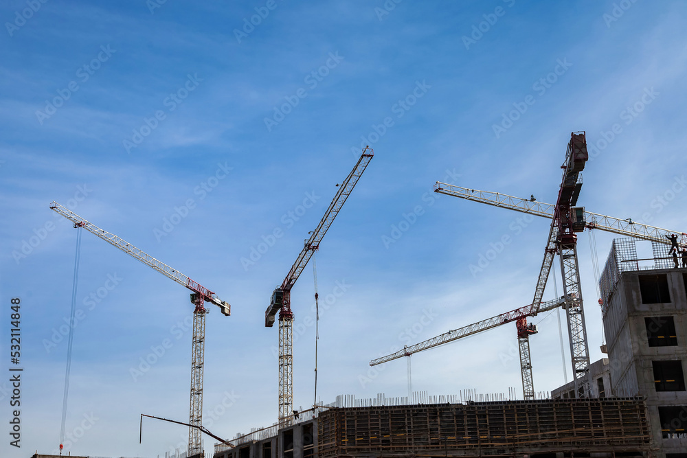 Construction cranes work on creation site against blue sky background. Bottom view of industrial crane. Concept of construction of apartment buildings and renovation of housing. Copy text space