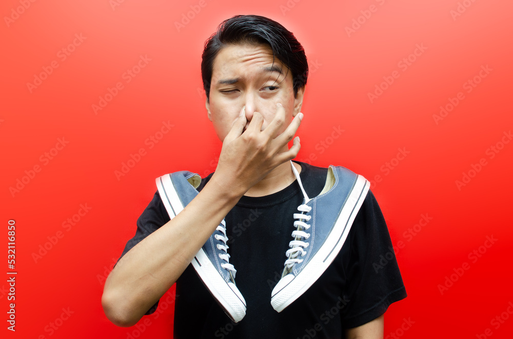 Disgusted asian man with black t-shirt holding a pair of stinky shoes isolated on red background. cover up the nose using hands to stop smelling bad. Disgusted asian man with black t-shirt holding a pair of stinky shoes isolated on red background. cover up the nose using hands to stop smelling bad.