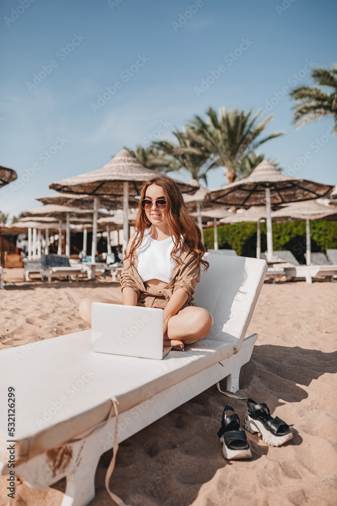 A cheerful blogger girl remotely works with a laptop on the beach with palm trees. A woman makes a video from a trip. Travels the world and works with social networks online. Remote modern profession