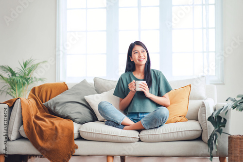 Happy young asian woman drinking coffee relaxing on sofa at home. Smiling female enjoying resting sitting on couch in modern living room.