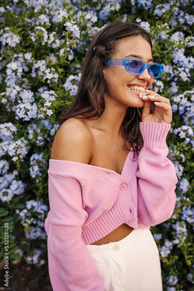 Cheerful young caucasian girl smiles teeth looking to side standing near tree with flowers. Brunette wears casual clothes, sunglasses. Lifestyle, different emotions, leisure concept.