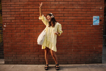 Cheerful young caucasian woman dancing listening to music in headphones outdoors. Brunette with closed eyes wears summer dress. Mood concept, use