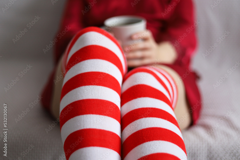 Woman in red white knee socks and silk nightie sitting on a sofa with tea cup in hands. Christmas holidays in home, warm drink in winter Woman in red white knee socks and silk nightie sitting on a sofa with tea cup in hands. Christmas holidays in home, warm drink in winter