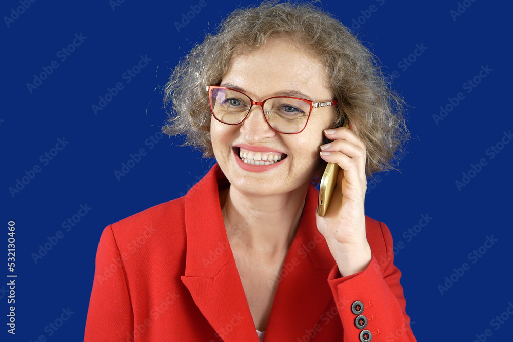 middle aged businesswoman in a red jacket holding a mobile phone. Female wearing eyeglasses looks at smartphone screen and smiling standing on a blue background with copy space,