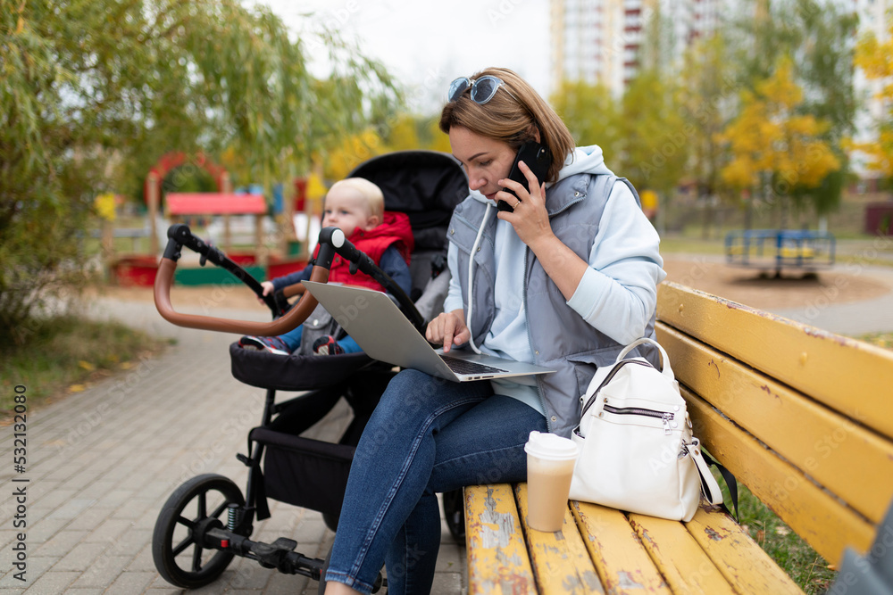 a young mother sitting on a bench next to a stroller with a child works online on a laptop and answers a phone call