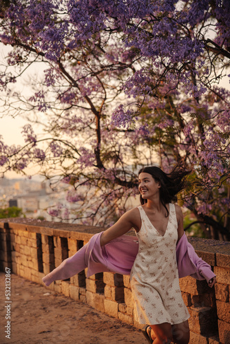 Energetic young caucasian girl in sundress enjoys walk on warm spring day. Brunette looks to side, runs among sakura trees. Relaxation concept © Look!