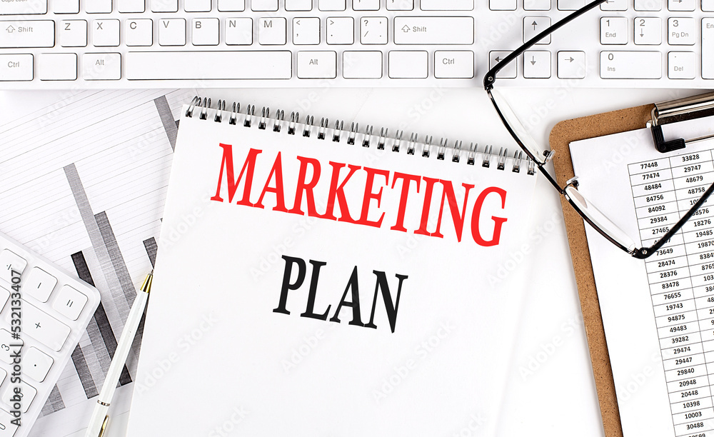 Text MARKETING PLAN on Office desk table with keyboard, notepad and analysis chart on white background. Text MARKETING PLAN on Office desk table with keyboard, notepad and analysis chart on white background.
