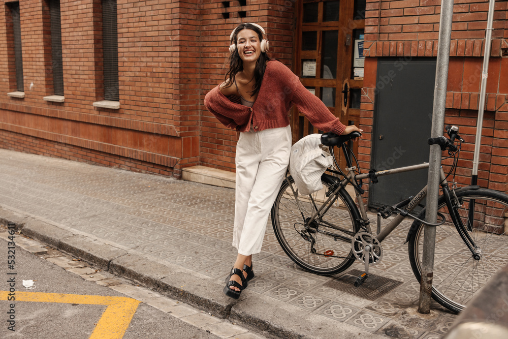Laughing full length young caucasian brunette wears headphones standing near bicycle on street. Woman in sweater and pants walks around city. Lifestyle concept.