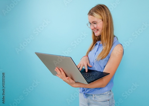 Smiling, confident, successful blonde woman in glasses holding and using laptop on blue isolated background. Freelancer