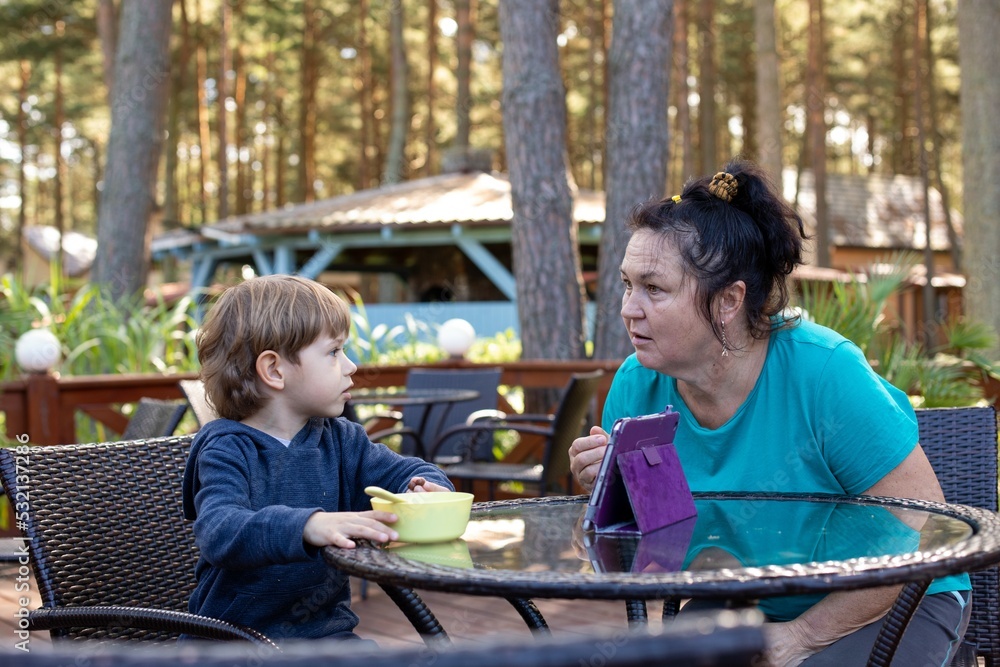 Little boy sit on chair at table on summer terrace and talk with mother. Kid of kindergarten age having dinner outdoor in forest. Communication with people, health resort, vacation, camp in forest Little boy sit on chair at table on summer terrace and talk with mother. Kid of kindergarten age having dinner outdoor in forest. Communication with people, health resort, vacation, camp in forest